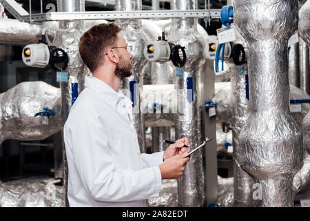 bearded engineer in white coat holding pen and clipboard near air compressed system Stock Photo