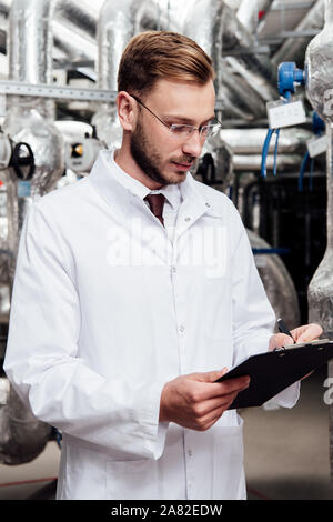 bearded engineer in white coat holding pen near clipboard and air compressed system Stock Photo
