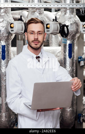 handsome bearded engineer in white coat and glasses holding laptop Stock Photo