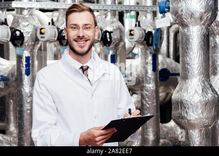 bearded and cheerful engineer in white coat holding pen and clipboard near air compressed system Stock Photo