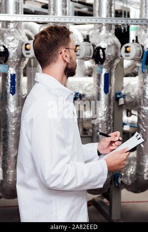 bearded engineer in white coat holding clipboard and pen near air compressed system Stock Photo