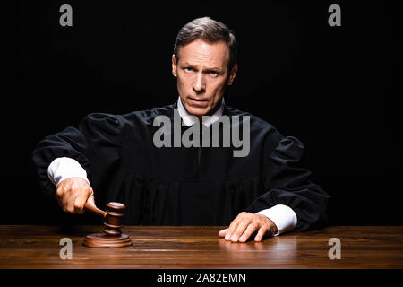 judge in judicial robe sitting at table and hitting with gavel isolated on black Stock Photo