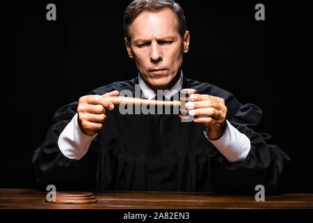 judge in judicial robe sitting at table and holding gavel isolated on black Stock Photo