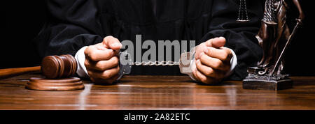 panoramic shot of judge in judicial robe sitting at table with handcuffs isolated on black Stock Photo