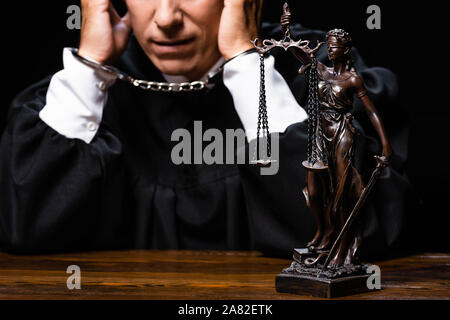 cropped view of judge in judicial robe sitting at table with handcuffs isolated on black Stock Photo