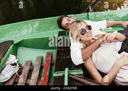 attractive girl in sunglasses smiling at camera while lying in boat near boyfriend Stock Photo