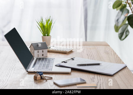 wooden desk with laptop, smartphone, house keys, clipboard with pen, house model and green plant Stock Photo