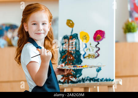 smiling redhead child standing near painting on canvas in art school Stock Photo