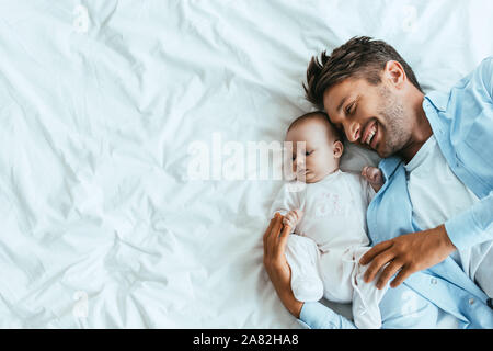 top view of happy father hugging adorable baby while lying on white bedding Stock Photo