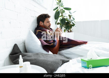 ill man with cup of hot drink lying in bed with napkins and nasal spray Stock Photo