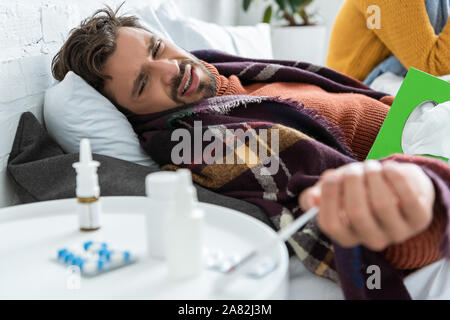 ill man in bed with pills, napkins and nasal spray in bedroom with woman Stock Photo