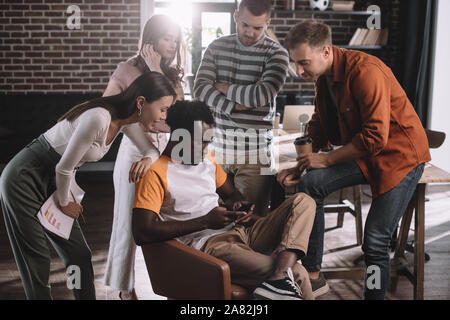 concentrated african american businessman using smartphone while sitting in armchair surrounded by young multicultural colleagues Stock Photo