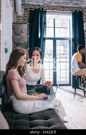 young businesswoman using laptop while sitting on sofa near smiling colleague Stock Photo