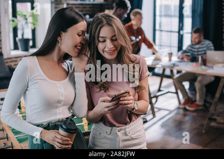 attractive, smiling businesswoman using smartphone near cheerful colleague holding coffee to go Stock Photo