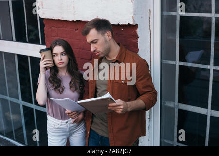 tired businesswoman holding coffee to go near head while standing near thoughtful colleague with documents Stock Photo