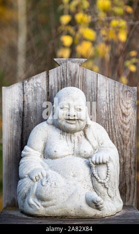 The Laughing Buddha, Budai, with his cloth sack stone sculpture with yellow fall colors in the background in autumn Stock Photo