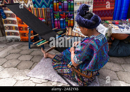A Mayan woman in traditional dress sitting on the ground and weaving on a backstrap loom in Santa Catarina Palopo, Guatemala.  Finished weavings hang Stock Photo