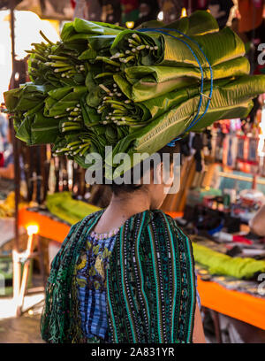 A young Mayan woman in traditional dress carries a bundle of banana leaves on her head in the market at Santiago Atitlan, Guatemala.  The leaves are s Stock Photo