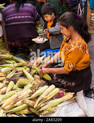 A woman husks corn at the market in Chichicastenango, Guatemala while her young son eats.  The boy appears to be about 4 years old. Stock Photo