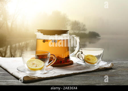 Jug of hot tea and cups on table near foggy pond Stock Photo