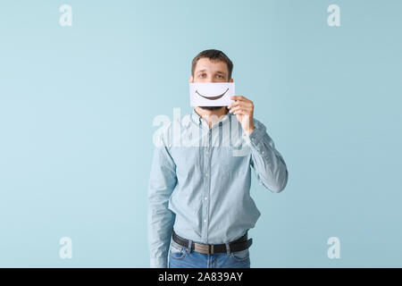 Man hiding mouth behind sheet of paper with drawn smile on color background Stock Photo