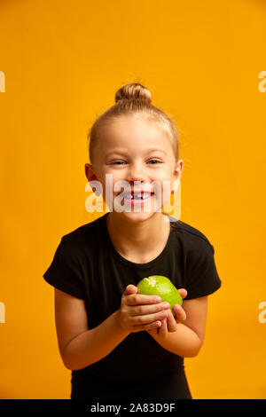 little dancer portrait on yellow background, portrait of a happy child smiling and showing off her first lost milk tooth in her hand a green apple Stock Photo