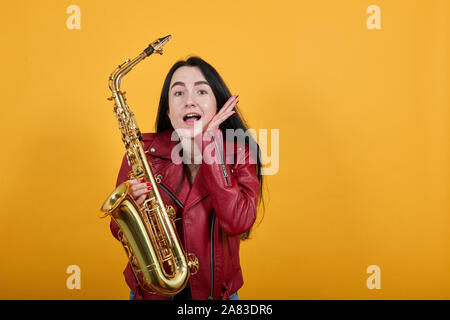 Shocked young woman keeping mouth open and saxophone, putting hands on cheeks Stock Photo