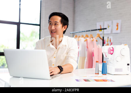 Contemporary confident Asian male entrepreneur and fashion designer in white shirt working on laptop with garment hanger on background Stock Photo