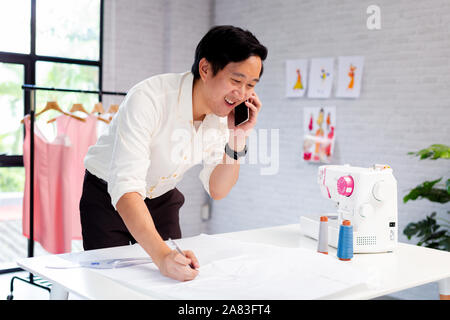 Asian businessman in white shirt smiling while discussing fashion project with partners on mobile phone in design studio with clothes on rack Stock Photo