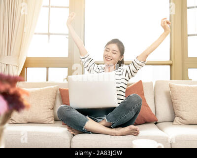 beautiful and happy young asian woman working from home sittng on couch using laptop computer, arms raised celebrating success Stock Photo