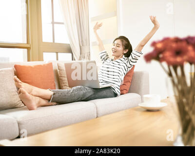 beautiful and happy young asian woman working from home sittng on couch using laptop computer, arms raised celebrating success