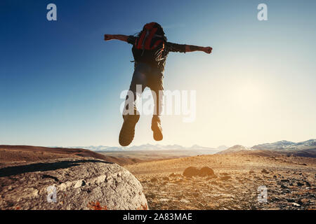 Hiker with backpack jumps from big rock against mountains and sunset Stock Photo