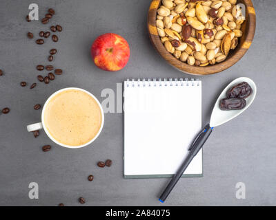Coffee break in the workplace. White cup of coffee, apple, mix of nuts in wooden bowl, notepad, pen on grey background, top view Stock Photo