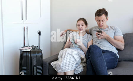 Young couple, man and woman choose budget hotel for rest using mobile phone. Woman is counting cash money. They are sitting on sofa in their flat. Stock Photo