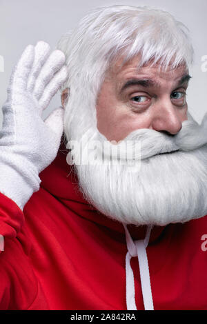 Portrait of a Santa Claus in sportsware. White-haired elderly man with white beard and moustache looking in camera? holds hand by ear and listens Stock Photo