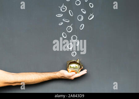 Concept of saving money with a gold colored piggy bank held in a hand in front of a blackboard with hand drawn coins Stock Photo
