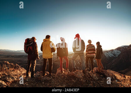 Group of six tourists backpackers or hikers stands against sunset light and mountains