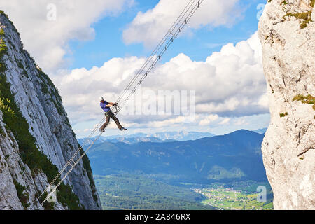 Man posing on Donnerkogel Intersport via ferrata (klettersteig) ladder, with his leg  hanging out, on a bright sunny day. Stock Photo