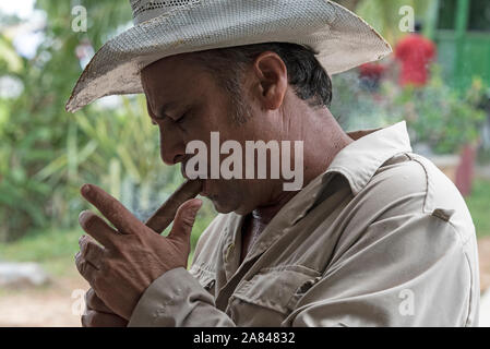 A Cuban farmer/ owner wearing a traditional Cuban hat lights his hand-rolled cigar using a lighter, in the Valle de Vinales, Pinar del Río Province, a Stock Photo