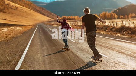 Young couple is riding on longboards by straight mountain road at sunset light Stock Photo