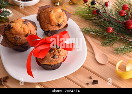 Chocolate chip muffin on white plate on wooden table decorated with Christmas objects with red bow. Horizontal composition. Elevated view. Stock Photo