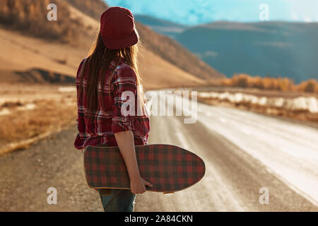 Pretty girl stands with longboard in hands at straight road against mountains and sunset