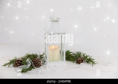 Christmas copy space with a lit candle inside a white lantern, pine branches, white organza ribbon and natural pine cones on a light wooden background Stock Photo