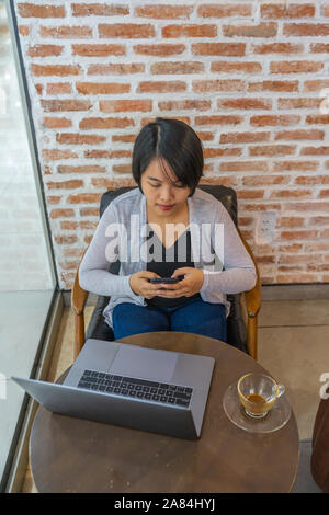 Asian woman looking at smartphone screen while sitting with laptop Stock Photo