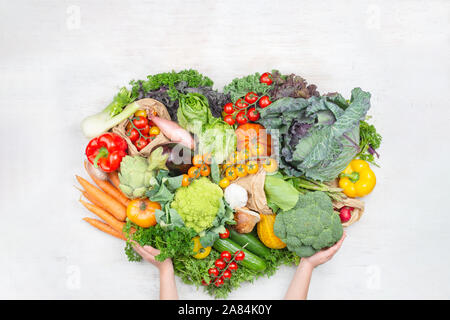 Healthy shopping. Child's hands holding rainbow vegetables in a shape of the heart on white wooden table tomatoes cabbage greens carrots broccoli, top view, on white wooden table, selective focus Stock Photo