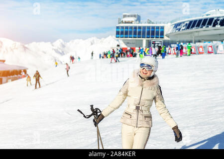Portrait of young adult beautiful happy caucasian woman smiling near lift station at alpine winter skiing resort. Girl in fashion ski suit, goggles Stock Photo