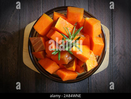 Top view of pieces of pumpkin with rosemary in a round glass plate on dark background. Raw vegetables. Stock Photo