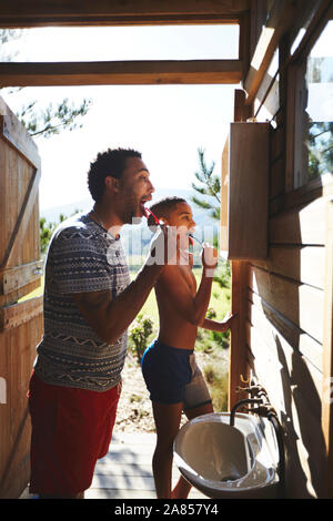 Father and son brushing teeth at sunny campsite bathroom mirror Stock Photo