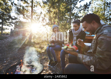 Happy friends eating at sunny campsite in woods Stock Photo