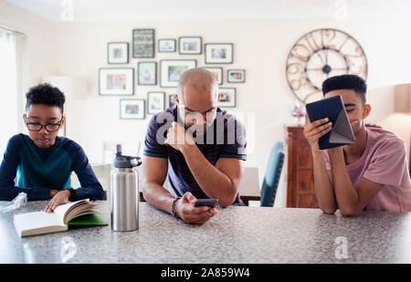 Father and sons using digital tablet, smart phone and reading book in kitchen